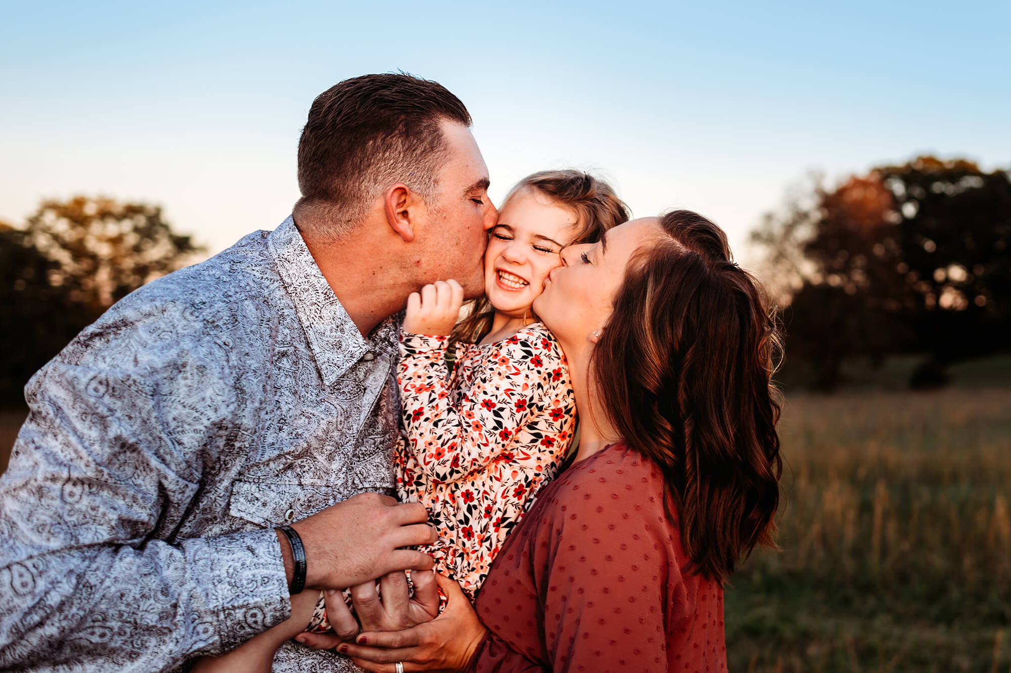Springfield MO family photographer captures toddler being kissed by dad and mom in open field at sunset