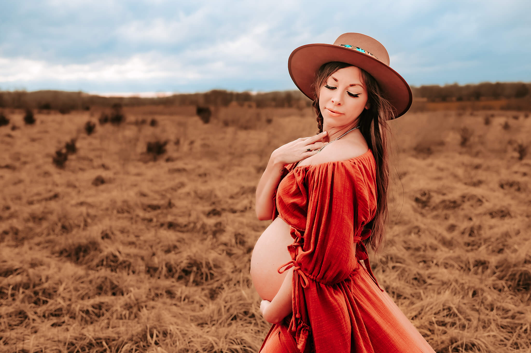 maternity photographer in Kansas City captures pregnant mom in field in red dress holding belly with storm rolling in
