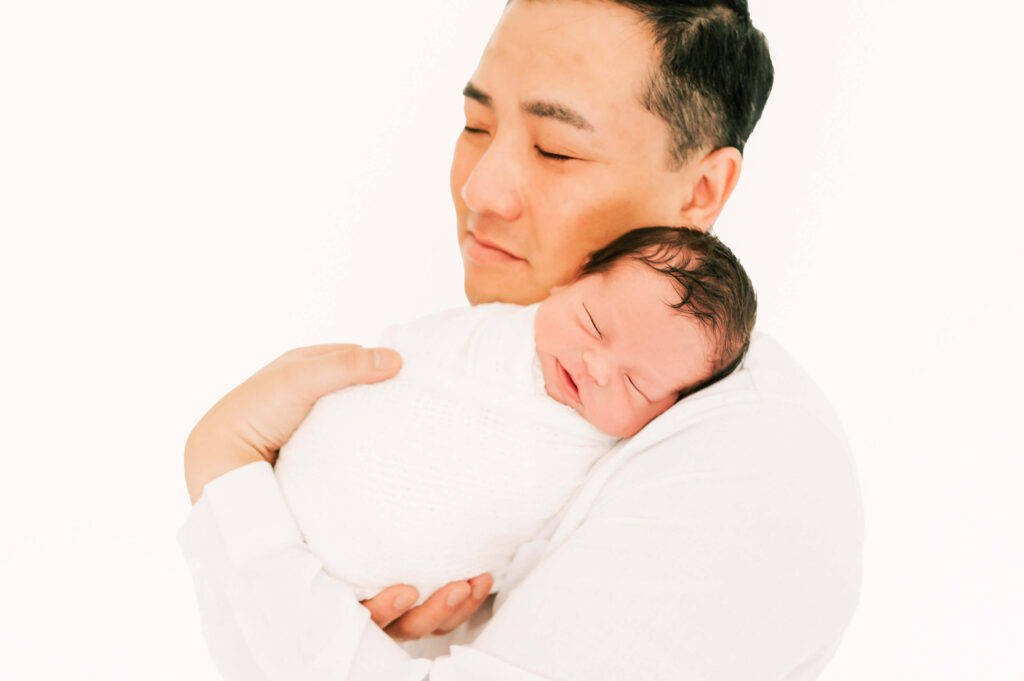 dad holding sleeping baby boy swaddled in white captured by The XO Photography